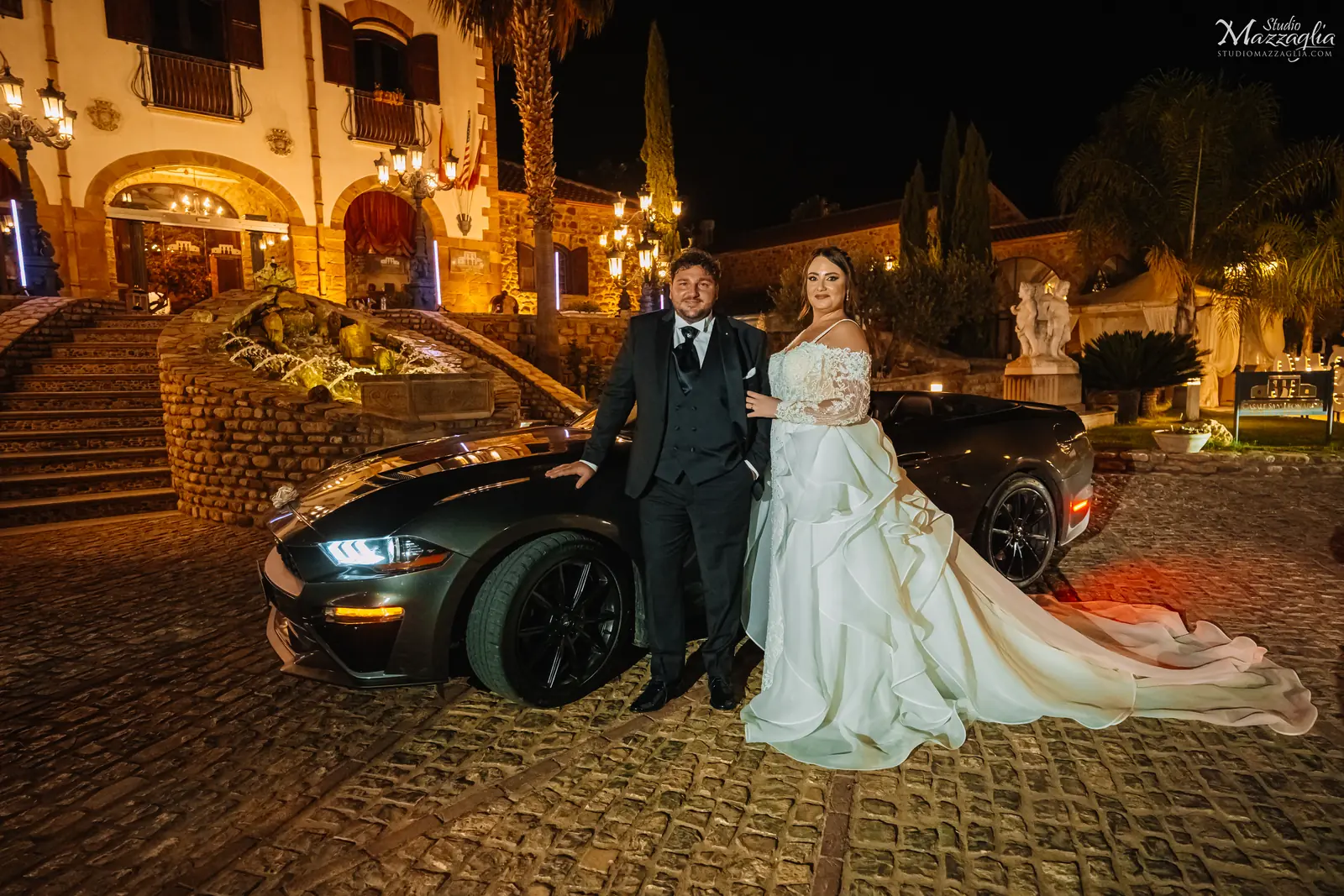 Sposi posano di notte accanto alla loro auto nuziale Mustang all'esterno illuminato del Casale San Leonardo durante il ricevimento.