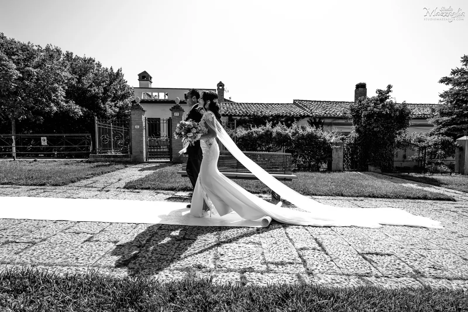 Il percorso verso il Sì Scatto in bianco e nero a figura intera che ritrae la sposa mentre percorre una passerella bianca all'aperto, accompagnata dal padre.