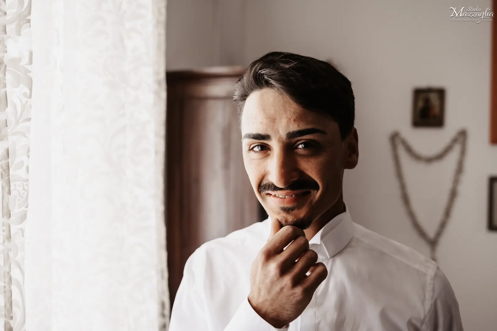 Ritratto dello sposo Ritratto ravvicinato di uno sposo che sorride, guardando in camera e toccandosi il mento con la mano destra. Indossa una camicia bianca abbottonata.