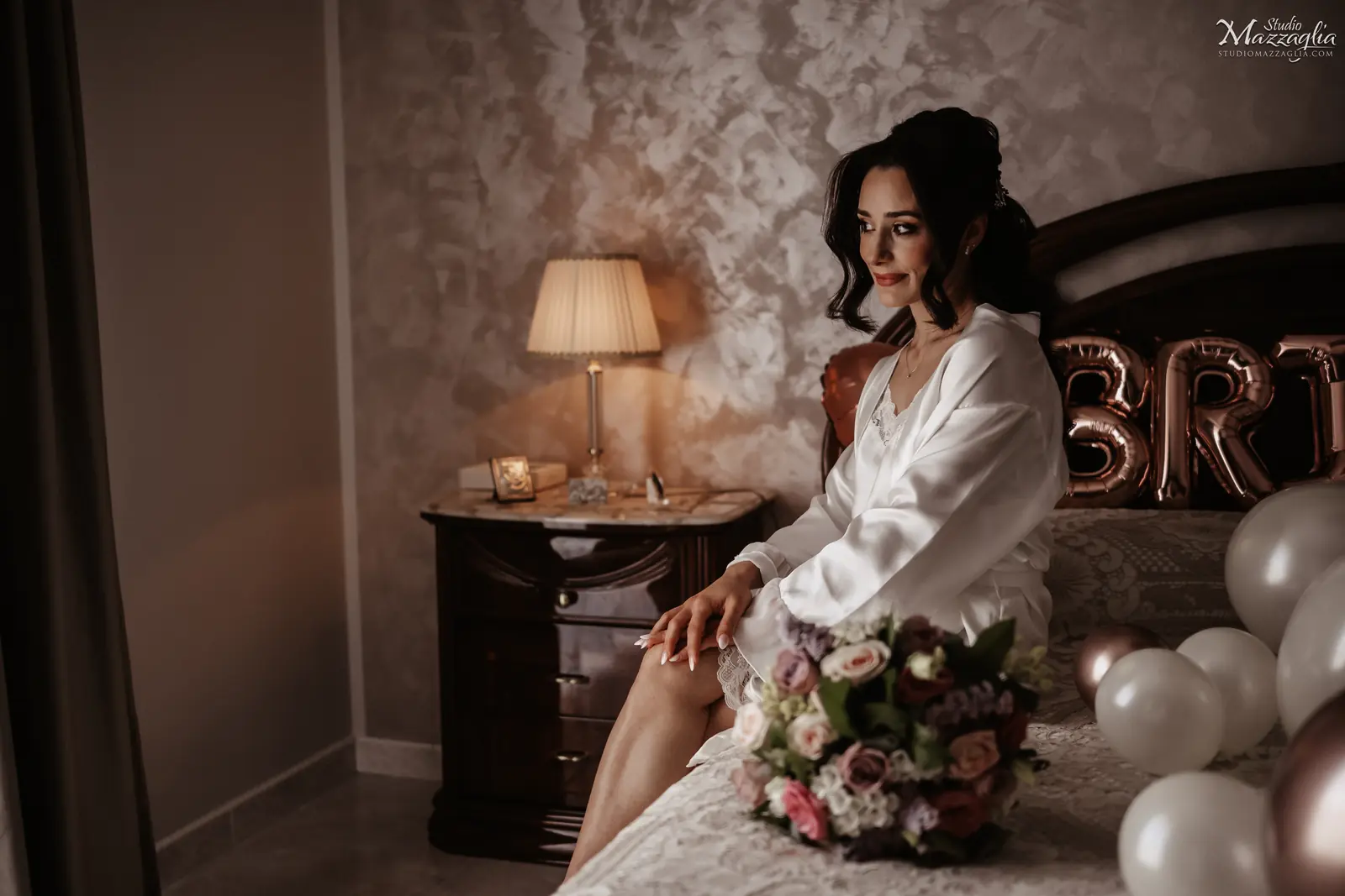 La sposa prima dell'abito La sposa, con indosso una vestaglia di seta bianca e un'acconciatura raccolta, siede sul bordo di un letto decorato con palloncini e la scritta "BRIDE".